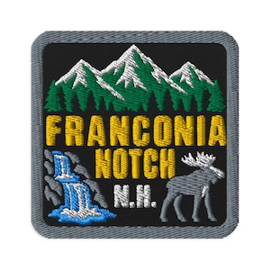 Può includere: Una toppa ricamata quadrata con bordo grigio su sfondo nero, raffigurante cime montuose bianche e verdi, una cascata blu, un alce grigio e il testo giallo che recita FRANCONIA NOTCH N.H. in un carattere grassetto senza grazie.