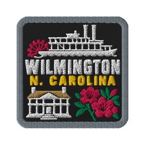 Puede incluir: Un parche bordado cuadrado con fondo negro y borde gris. Presenta un barco de vapor blanco, el texto "WILMINGTON N. CAROLINA" en blanco y amarillo, un edificio blanco y un grupo de flores rosas con hojas verdes.