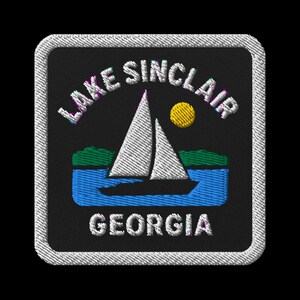 Może przedstawiać: Haftowana naszywka przedstawiająca żaglówkę na jeziorze Sinclair w stanie Georgia. Projekt zawiera białą żaglówkę, niebieską wodę, zielony ląd i żółte słońce. Tekst "LAKE SINCLAIR GEORGIA" jest haftowany na biało.