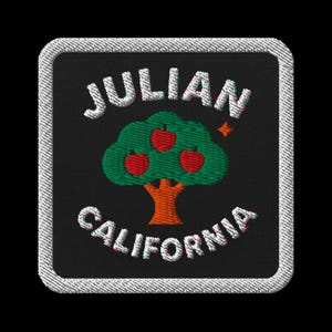 Peut inclure: Écusson brodé avec les mots "JULIAN CALIFORNIA" en blanc, encadrant un arbre vert avec trois pommes rouges. L'écusson a un fond noir et une bordure blanche cousue.