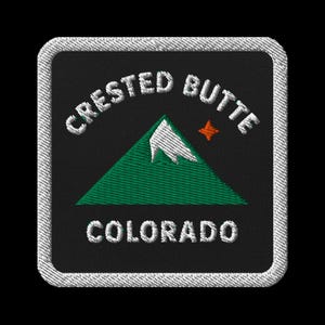 May include: Embroidered patch featuring the words "CRESTED BUTTE COLORADO" in white, arched above and below a green mountain with a white snow-capped peak and a small orange star. The patch has a black background and a white stitched border.