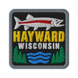 Könnte beinhalten: Ein quadratischer gestickter Aufnäher mit grauem Rand auf schwarzem Hintergrund. Er zeigt einen weißen Fisch mit rotem Streifen, den gelben Schriftzug HAYWARD, den weißen Schriftzug WISCONSIN, grüne Tannen und blaue Wellenlinien.