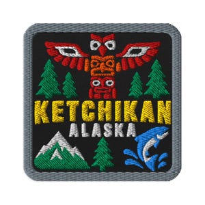 May include: A square embroidered patch with a black background featuring a red totem pole, green pine trees, and a blue jumping fish. The text KETCHIKAN is stitched in yellow, and ALASKA is in white. A mountain peak is depicted in white and green.