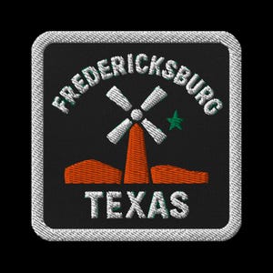 Pode incluir: Patch bordado com as palavras "FREDERICKSBURG TEXAS" em branco, sobre um desenho de moinho de vento e estrela. O moinho de vento é branco com uma torre laranja. O patch tem um fundo preto e uma borda branca costurada.