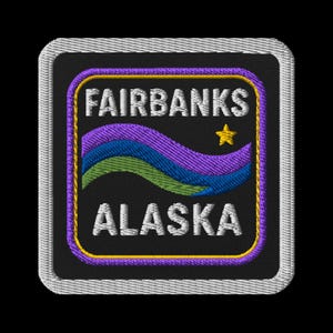 Pode incluir: Patch bordado com as palavras "FAIRBANKS ALASKA" em branco, acima e abaixo de um design ondulado em roxo, azul e verde. Uma estrela dourada está à direita da palavra "FAIRBANKS". O patch tem um fundo preto e uma borda prateada.
