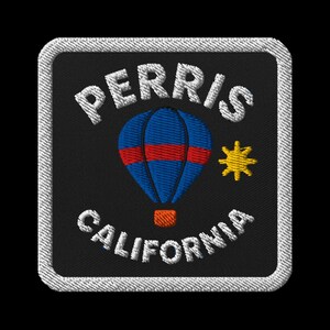Puede incluir: Parche bordado con el texto "PERRIS CALIFORNIA" en blanco, arqueado alrededor de un globo aerostático azul con rayas rojas y una cesta naranja. Una estrella amarilla está a la derecha del globo. El parche tiene un borde blanco.