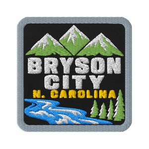 Op de afbeelding: Een vierkante geborduurde patch met een zwarte achtergrond met groene bergen met witte besneeuwde toppen, een blauwe rivier en groene dennenbomen. De tekst luidt BRYSON CITY in het wit en N. CAROLINA in het geel met een grijze rand.