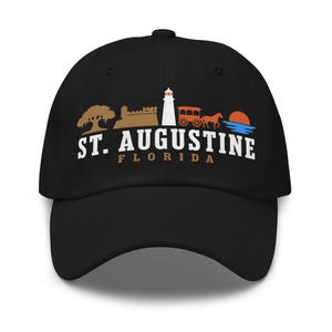 Könnte beinhalten: Eine schwarze Baseballkappe mit einer Grafik von St. Augustine, Florida. Das Design zeigt einen Baum, eine Festung, einen Leuchtturm, eine Pferdekutsche und einen Sonnenuntergang über dem Wasser, mit dem Text ST. AUGUSTINE FLORIDA in Weiß und Braun.
