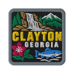 Könnte beinhalten: Ein quadratischer gestickter Aufnäher mit schwarzem Hintergrund und grauem Rand. Er zeigt einen weißen Wasserfall, grüne Kiefern und einen Berg. Der Text CLAYTON GEORGIA ist in Gelb und Weiß gestickt. Unten sind eine rosa Blume und ein blauer Fisch.