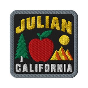 Puede incluir: Un parche bordado cuadrado con fondo negro y borde gris. El texto JULIAN está en amarillo en la parte superior y CALIFORNIA en blanco en la parte inferior. Una manzana roja, un pino verde, un sol amarillo y montañas amarillas están centrados.