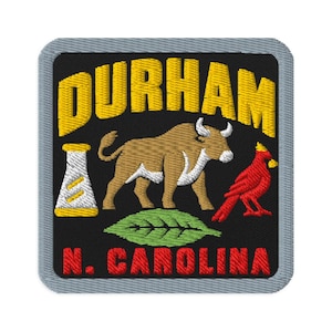 Puede incluir: Un parche bordado cuadrado con borde gris sobre fondo negro. Presenta el texto amarillo DURHAM sobre un toro marrón, un matraz de laboratorio blanco, un pájaro cardenal rojo, una hoja verde y el texto rojo N. CAROLINA debajo.