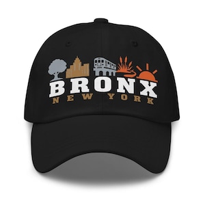Puede incluir: Una gorra de béisbol negra con un diseño gráfico que muestra un árbol, un edificio, un tren subterráneo, juncos y un sol. Debajo de los iconos, el texto BRONX en letras blancas grandes y NEW YORK en letras color canela está centrado.