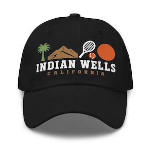 Puede incluir: Una gorra de béisbol negra con un gráfico que incluye una palmera verde, una montaña marrón, una raqueta de tenis blanca, una pequeña pelota de tenis naranja y un gran círculo naranja. El texto dice INDIAN WELLS CALIFORNIA en letras.