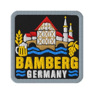 May include: A square embroidered patch with a grey border on a black background. It features a white and red timber-framed building, a bridge, a beer mug, and hops. The text reads BAMBERG in yellow and GERMANY in white with blue wave accents.