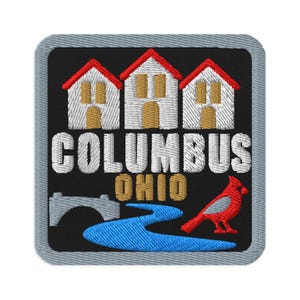 Pode incluir: Um emblema bordado quadrado com fundo preto apresentando três casas brancas com telhados vermelhos, o texto COLUMBUS OHIO em branco e dourado, uma ponte de pedra cinza, um rio azul sinuoso e um pássaro cardeal vermelho, tudo contornado por uma moldura cinza.