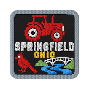 Puede incluir: Un parche bordado cuadrado con borde gris sobre fondo negro. Presenta un tractor rojo, el texto blanco SPRINGFIELD, el texto amarillo OHIO, un pájaro cardenal rojo, un puente arqueado blanco, un río azul y colinas verdes.