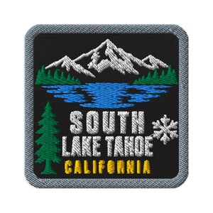 May include: A square embroidered patch with a black background and a gray border. It features a white mountain range above a blue lake, green pine trees, a white snowflake, and the white text SOUTH LAKE TAHOE with yellow text CALIFORNIA.