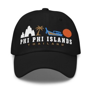 May include: A black baseball cap featuring a graphic design of white limestone cliffs, a brown palm tree, a blue long-tail boat, and an orange sun. Below the graphic, the text reads PHI PHI ISLANDS in white and THAILAND in brown lettering.