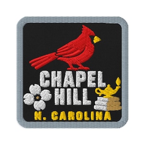 Può includere: Una toppa ricamata quadrata con sfondo nero e bordo grigio. Presenta un uccello cardinale rosso, un fiore di corniolo bianco e una lampada a olio gialla. Il testo recita CHAPEL HILL in bianco e N. CAROLINA in ricamo giallo.