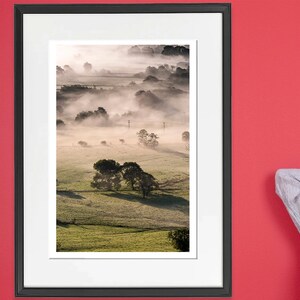 May include: A framed photograph of a misty landscape with rolling hills and trees. The photograph is in a black frame with a white mount.