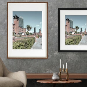 May include: Two framed photographs of a city street scene with brick buildings, palm trees, and a blue sky. The frames are brown wood and black.