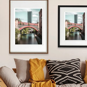 May include: Two framed photographs of a red bridge over a canal in a city. The bridge is made of brick and has a tall building in the background. The photos are in different sizes and frames.