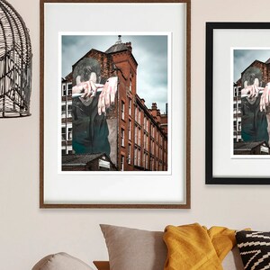 Op de afbeelding: Twee ingelijste foto's van een muurschildering met straatkunst die het gezicht en de handen van een persoon laat zien die uit een bakstenen gebouw komen. De muurschildering is geschilderd in tinten grijs, bruin en rood.