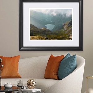 May include: A framed photograph of a mountain lake surrounded by green hills and clouds. The photo is in a black frame with a white mount.