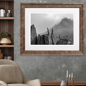 Op de afbeelding: Een zwart-witfoto van een rotsachtig berglandschap met mist op de achtergrond. De foto is ingelijst in een rustiek houten frame.