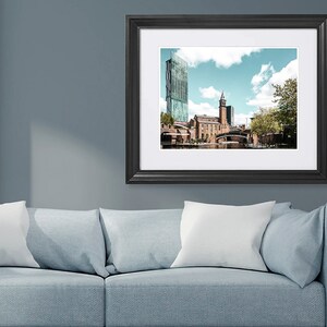 May include: A framed photograph of a city skyline with a canal in the foreground. The buildings are a mix of modern and traditional architecture, with a tall glass building on the left and a brick building with a clock tower on the right. The canal is lined with trees and there is a blue sky with white clouds in the background.
