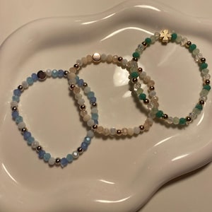Könnte beinhalten: Drei Armbänder mit Perlen in Blau, Pfirsich und Grün. Jedes Armband hat kleine, facettierte Perlen und metallische Akzente. Ein Armband hat einen goldfarbenen vierblättrigen Kleeblatt-Anhänger. Die Armbänder werden auf einer weißen Schale mit gewelltem Rand präsentiert.