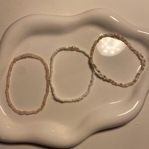 Könnte beinhalten: Drei Armbänder aus Perlen in Rosa-, Weiß- und Goldtönen liegen auf einer weißen, organisch geformten Schale. Die Armbänder bestehen aus kleinen, runden Perlen, die zusammen aufgefädelt sind und eine zarte und feminine Ästhetik erzeugen. Die Schale bietet einen sauberen, neutralen Hintergrund.