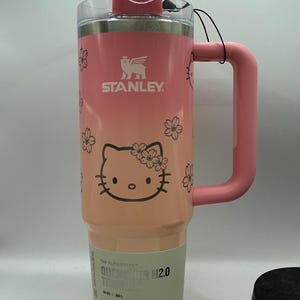 May include: A pink Stanley Quencher H2.0 tumbler with a 40 oz capacity, featuring a Hello Kitty graphic with cherry blossoms, a pink handle, a clear lid with a pink cap, and a label stating The FlowState Quencher H2.0 Tumbler 40 oz 1.18 L.