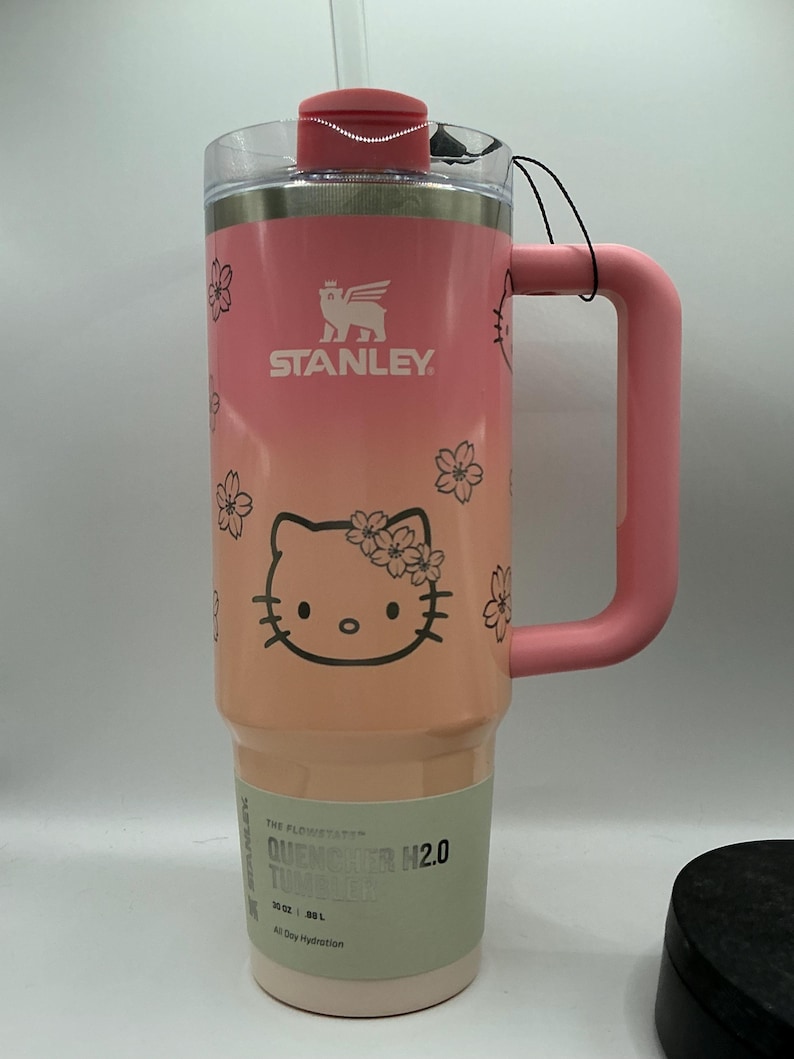 May include: A pink Stanley Quencher H2.0 tumbler with a 40 oz capacity, featuring a Hello Kitty graphic with cherry blossoms, a pink handle, a clear lid with a pink cap, and a label stating The FlowState Quencher H2.0 Tumbler 40 oz 1.18 L.