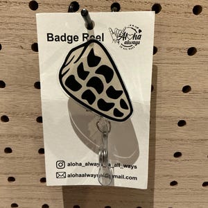 May include: Badge reel with a seashell design. The badge reel features a clear plastic retractable cord and a white card with the text "Badge Reel" and social media information. The seashell is white with black spots.