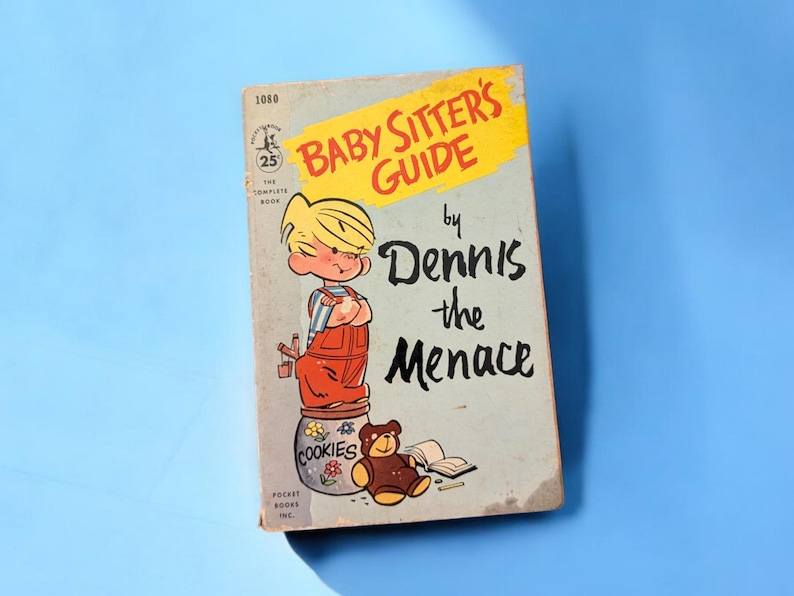May include: A vintage paperback book titled "Baby Sitter's Guide by Dennis the Menace". The cover features a cartoon illustration of Dennis the Menace standing on a cookie jar with a teddy bear beside him.