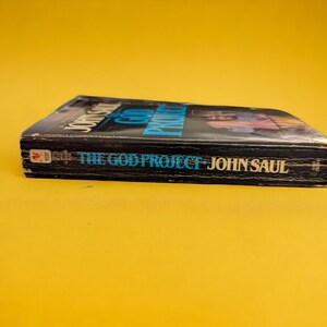 May include: A hardcover book titled "THE GOD PROJECT" by John Saul. The book has a black cover with blue and white text. The book is set against a yellow background.