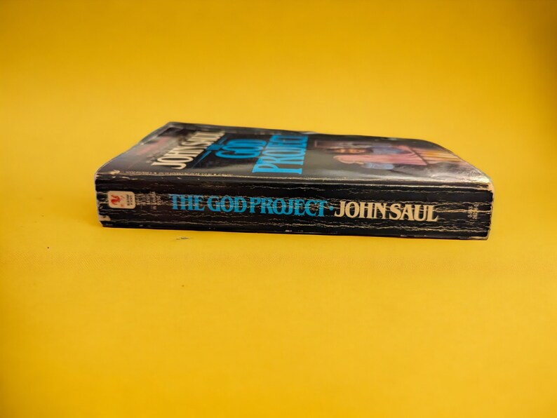 May include: A hardcover book titled "THE GOD PROJECT" by John Saul. The book has a black cover with blue and white text. The book is set against a yellow background.