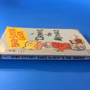 May include: A vintage paperback book titled "Baby Sitter's Guide by Dennis the Menace". The cover features a cartoon illustration of Dennis the Menace holding a jar of cookies.
