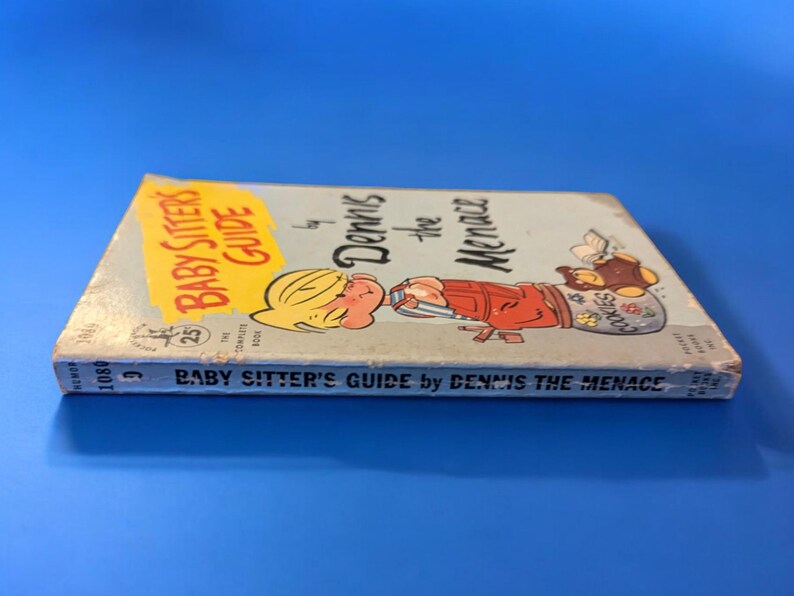 May include: A vintage paperback book titled "Baby Sitter's Guide by Dennis the Menace". The cover features a cartoon illustration of Dennis the Menace holding a jar of cookies.