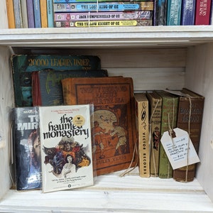 May include: A wooden crate shelf filled with vintage books. Titles include "20,000 Leagues Under the Sea" and "The Haunted Monastery." The books vary in color, with some bound in green and brown. A price tag is attached to a bundle of books.