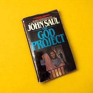 May include: A paperback book titled "The God Project" by John Saul. The cover features a dark background with the title in large, blue letters. A person is shown in the illustration, holding a pink blanket, with a shadowy figure in the background.