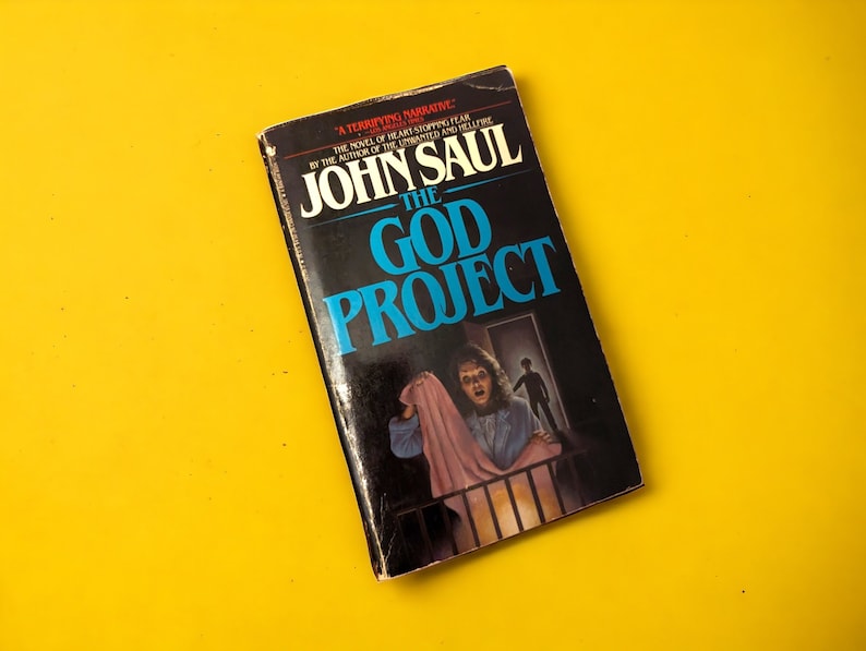 May include: A paperback book titled "The God Project" by John Saul. The cover features a dark background with the title in large, blue letters. A person is shown in the illustration, holding a pink blanket, with a shadowy figure in the background.