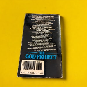 May include: A paperback book titled "The God Project" with a black cover and white text. The back cover features a synopsis of the story, with text about missing children and a town in Massachusetts. The book is set against a yellow background.