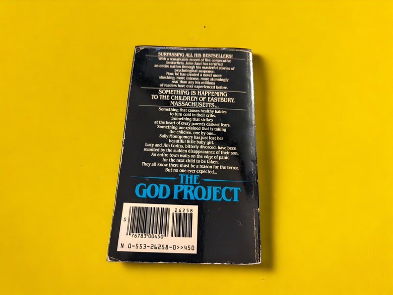 May include: A paperback book titled "The God Project" with a black cover and white text. The back cover features a synopsis of the story, with text about missing children and a town in Massachusetts. The book is set against a yellow background.