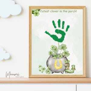 Op de afbeelding: Een groene handafdruk wordt getoond die uit een pot met een gouden hoefijzer groeit. De pot is gevuld met groene klavers en er is een groene kabouterlaars en -hoed naast de pot. De tekst "Cutest clover in the porch!" staat bovenaan de afbeelding.
