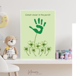 Op de afbeelding: Een groene handafdruk met de tekst "Cutest clover in the porch!" erboven. De handafdruk bevindt zich op een groene achtergrond met groene vierbladige klavers onderaan.