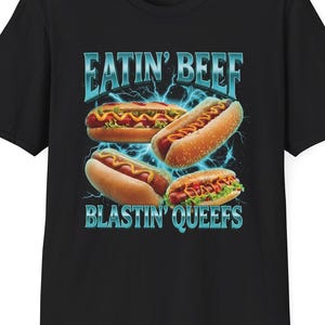 May include: A black short-sleeve t-shirt featuring a graphic print of four hot dogs arranged in a cluster. The text EATIN' BEEF appears above the hot dogs, and BLASTIN' QUEEFS appears below, both in a metallic blue font against a lightning background.