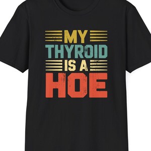 May include: Black t-shirt with the text "MY THYROID IS A HOE" in a retro, distressed font. The text is in yellow, teal, and orange, with horizontal lines above and below the words. The shirt is a classic crew neck style.