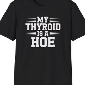 May include: Black t-shirt with the white text "MY THYROID IS A HOE" in a bold, sans-serif font. The text is arranged in three lines, with the words "MY" and "IS A" separated by horizontal lines. The shirt has a crew neck and short sleeves.